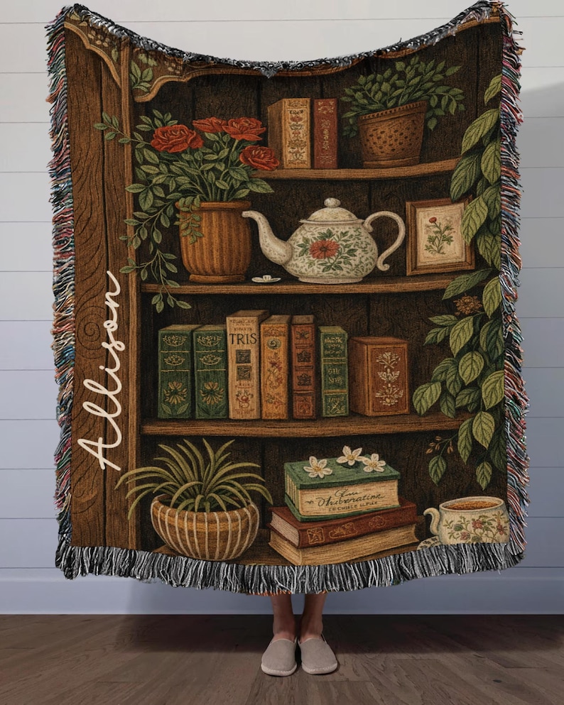 May include: A woven throw blanket featuring a bookshelf design with books, plants, and a teapot. The blanket has a brown background with the name "Allison" written vertically on the left side. The edges are fringed with multiple colors.