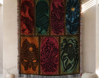 May include: A woven throw blanket featuring six rectangular panels with intricate illustrations including a bird, a dagger, a flower, a mask, a campfire, and a spider, all rendered in vibrant red, green, blue, and orange tones with fringe edges.