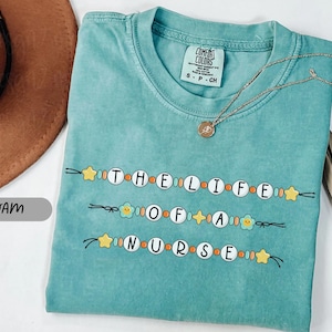 May include: Seafoam green t-shirt with the phrase "THE LIFE OF A NURSE" in a beaded design. The shirt has a crew neck and short sleeves. A gold necklace with a pendant is draped on the shirt.