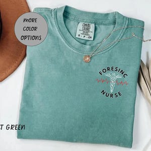 May include: Light green t-shirt with the text "FORENSIC NURSE" and a medical symbol. The shirt has a crew neck and short sleeves. A gold necklace is draped across the shirt. The words "MORE COLOR OPTIONS" are in a circle.