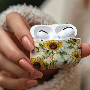 May include: White wireless earbuds in a decorative case featuring a sunflower design. The case has a cream background with yellow sunflowers, green leaves, and gold accents. The earbuds are open and ready to use.