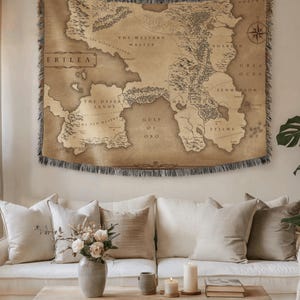 May include: A rectangular woven tapestry featuring a tan and brown fantasy map of Erilea, including labels like The Western Wastes, The Desert Lands, Gulf of Oro, and Eyllwe. The map has a compass rose and fringed edges on the top and bottom sides.