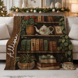 May include: A cozy throw blanket featuring a detailed illustration of a wooden bookshelf filled with books, a teapot, and potted plants. The blanket has a warm, inviting color palette with the name "Allison" written in script.