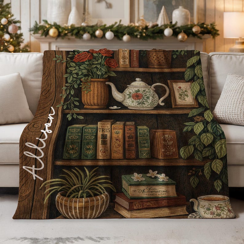 May include: A cozy throw blanket featuring a detailed illustration of a wooden bookshelf filled with books, a teapot, and potted plants. The blanket has a warm, inviting color palette with the name "Allison" written in script.