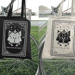 May include: Black and off-white tote bags with a witch-themed design. Each bag features a rectangular graphic with a group of witches, bats, and the text "GIRLS WILL BE GIRLS". The bags are hanging on a bicycle.
