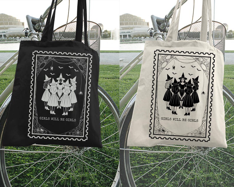 May include: Black and off-white tote bags with a witch-themed design. Each bag features a rectangular graphic with a group of witches, bats, and the text "GIRLS WILL BE GIRLS". The bags are hanging on a bicycle.