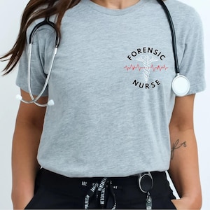 May include: A heather gray short-sleeve t-shirt featuring a black graphic on the chest that reads FORENSIC NURSE with a white caduceus symbol and a red heartbeat line. A black stethoscope is draped around the neck of the shirt.