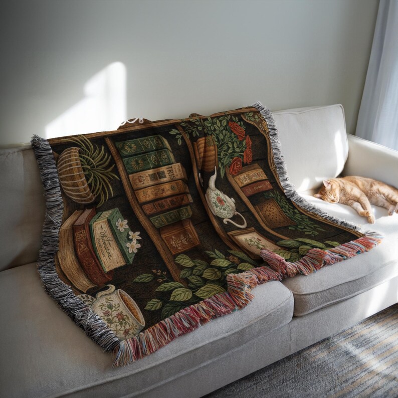 May include: A woven throw blanket featuring a bookshelf design with books, plants, and a teapot. The blanket has a dark brown background with colorful fringe along the edges. The blanket is draped over a white sofa.