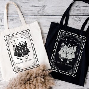 May include: Two tote bags, one cream and one black, with a witch-themed design. Each bag features a framed illustration of five witches in dresses and hats, with the text "GIRLS WILL BE GIRLS". The design includes spiderwebs and bats.