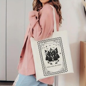 May include: Off-white tote bag featuring a black and white illustration of four witches in a stamp-like design. The text "GIRLS WILL BE GIRLS" is below the witches. The bag has a simple, rectangular shape.