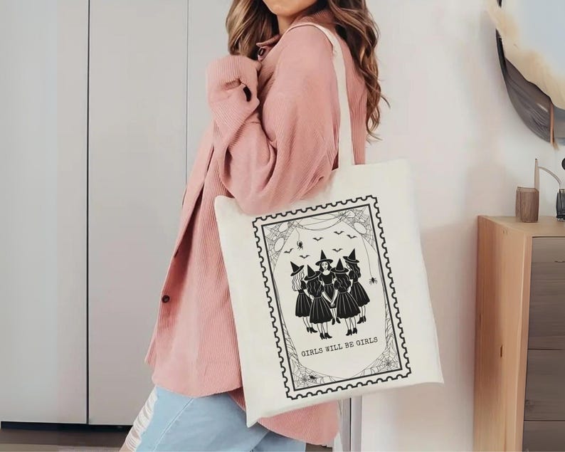 May include: Off-white tote bag featuring a black and white illustration of four witches in a stamp-like design. The text "GIRLS WILL BE GIRLS" is below the witches. The bag has a simple, rectangular shape.