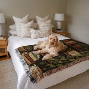May include: A tan dog rests on a bed covered with a decorative throw blanket. The throw features a dark background with images of books, plants, and a teacup. The bed has white linens and several pillows. Two lamps sit on the nightstands.