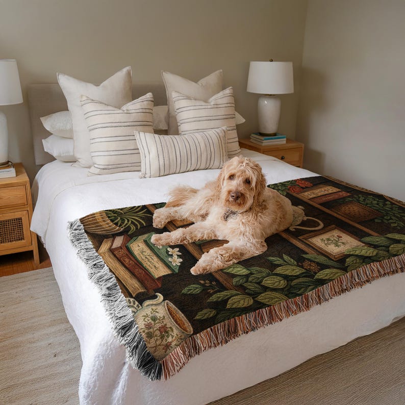 May include: A tan dog rests on a bed covered with a decorative throw blanket. The throw features a dark background with images of books, plants, and a teacup. The bed has white linens and several pillows. Two lamps sit on the nightstands.