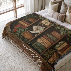 May include: A woven throw blanket featuring a bookshelf design with books, a teapot, and flowers. The blanket has a brown and black color scheme with the name "Allison" in white script on the side. The blanket is fringed.