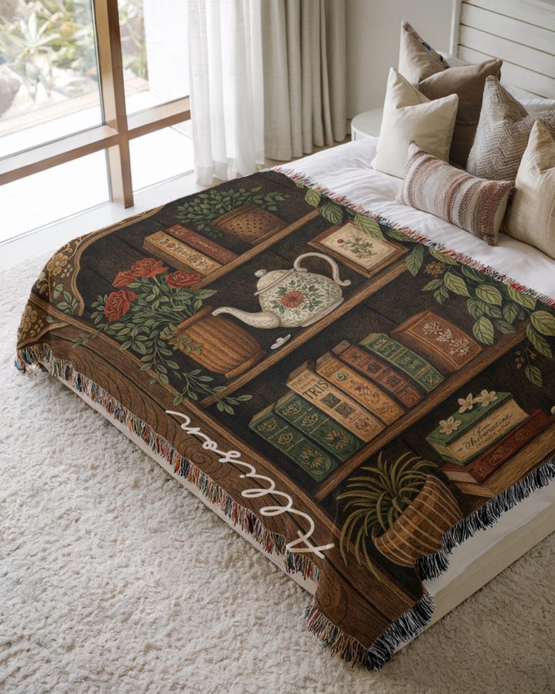 May include: A woven throw blanket featuring a bookshelf design with books, a teapot, and flowers. The blanket has a brown and black color scheme with the name "Allison" in white script on the side. The blanket is fringed.