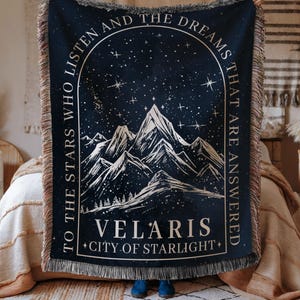 May include: A dark blue woven throw blanket featuring a white mountain range graphic under a starry sky. The text reads: TO THE STARS WHO LISTEN AND THE DREAMS THAT ARE ANSWERED, VELARIS, CITY OF STARLIGHT. The blanket has a fringed edge on all sides.