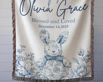 May include: A white woven throw blanket with a blue floral bunny design and the name "Olivia Grace". The blanket has a blue bow at the top and the text "Blessed and Loved" and "December 14, 2025". The edges are fringed.