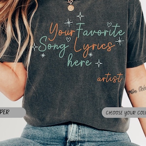 May include: A charcoal gray short-sleeve t-shirt featuring the text "Your Favorite Song Lyrics here" in a mix of orange and teal cursive and print fonts, accented with small white star and heart icons, with the word "artist" in orange below.