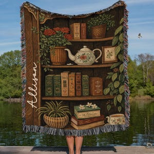 May include: A woven throw blanket featuring a detailed illustration of a bookshelf filled with books, plants, and a teapot. The design includes the name "Allison" in script. The blanket has a fringed edge and is set against a lake backdrop.