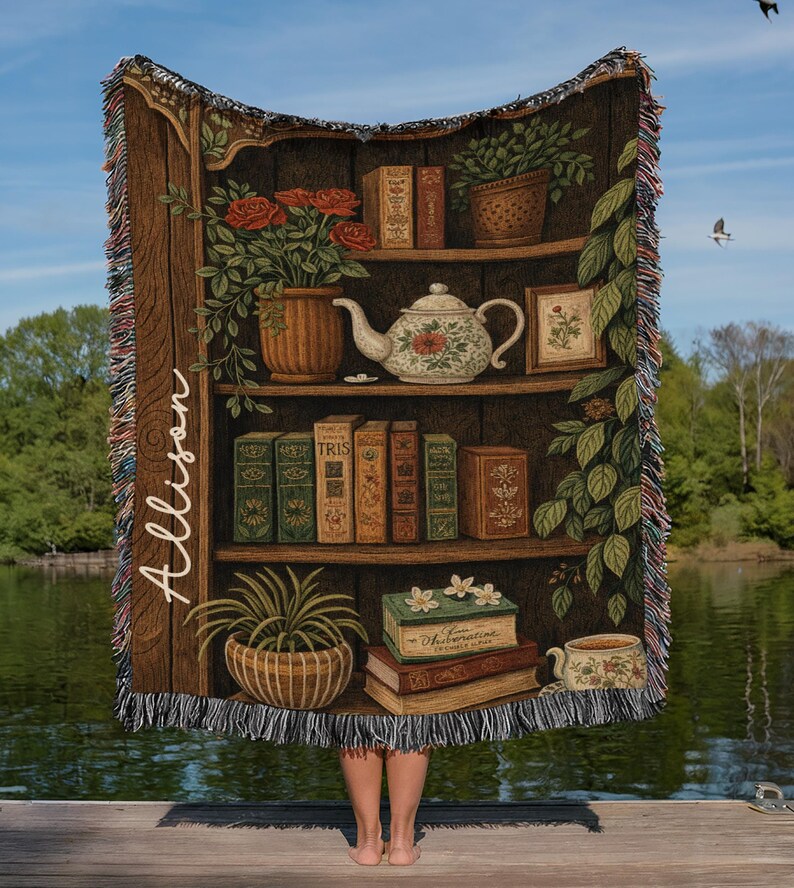 May include: A woven throw blanket featuring a detailed illustration of a bookshelf filled with books, plants, and a teapot. The design includes the name "Allison" in script. The blanket has a fringed edge and is set against a lake backdrop.