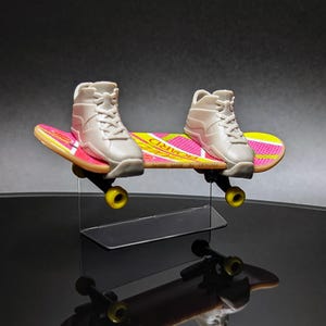Könnte beinhalten: Ein Miniatur-Skateboard mit rosa und gelbem Design, mit zwei grauen High-Top-Sneakern. Das Skateboard hat gelbe Rollen und wird auf einem transparenten Ständer ausgestellt. Der Text "BOARD MADE IN VIETNAM" ist auf dem Board sichtbar.