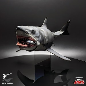 May include: A detailed, grey and white shark figurine with an open mouth, revealing teeth and a small object. The shark has a realistic design and is displayed on a stand. The text "DESIGNED TO FIT NECA JAWS" is visible.