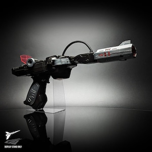 May include: A black and silver toy blaster with a pistol grip, a long barrel, and a curved black cable connecting the two sections. The device features red accents, a small red translucent sight, and is mounted on a clear acrylic display stand.