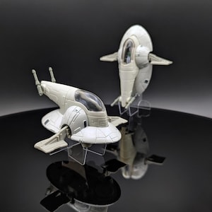 Könnte beinhalten: Zwei hellgraue Raumschiffmodelle werden auf transparenten Ständern vor einem dunklen Hintergrund ausgestellt. Die Schiffe haben ein unverwechselbares Design mit Flügeln und einem Cockpit. Eines befindet sich im Vordergrund, das andere dahinter.