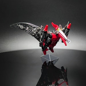 Op de afbeelding: Een rode, zwarte en zilveren speelgoedrobot in de vorm van een pterodactyl. Het speelgoed staat op een heldere standaard, met zijn vleugels gespreid. De robot heeft een gedetailleerd ontwerp, met zichtbare mechanische onderdelen en een heldere snavel.