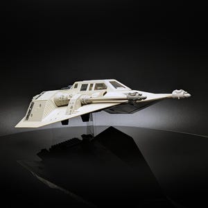 May include: A cream-coloured model of a Snowspeeder starfighter, a vehicle from a science fiction film. The model has a detailed design with wings, a cockpit, and gun attachments. The starfighter is set against a dark background.