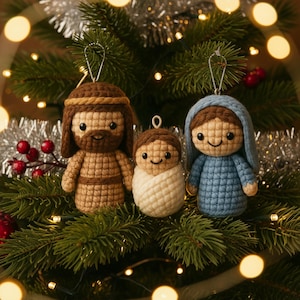 May include: Crocheted Christmas ornaments depicting the Nativity scene. The set includes figures of Joseph, baby Jesus, and Mary, each with smiling faces. The ornaments are brown, blue, and white, and are hanging on a Christmas tree.