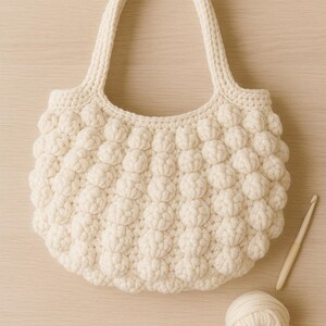 May include: Cream-colored crochet tote bag with a textured, puffy design. The bag has a rounded shape, two handles, and the text "Cloud Puff Tote" at the bottom. A ball of yarn and a crochet hook are next to the bag.
