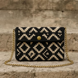 Może przedstawiać: Czarna i kremowa szydełkowa torba na ramię z geometrycznym wzorem. Torba posiada złoty łańcuszek i złote zapięcie na guzik. Torba leży na kamiennej powierzchni.