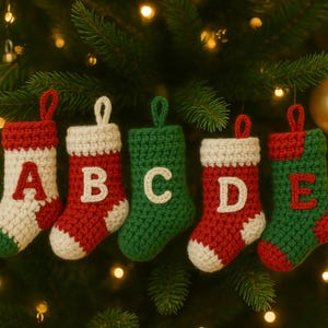 Op de afbeelding: Vijf gehaakte kerstsokornamenten in rood, wit en groen, elk met een andere letter: A, B, C, D en E. De sokken hangen aan een kerstboom met gouden ornamenten en lichtjes.