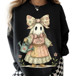 May include: Black sweatshirt featuring a whimsical illustration of a ghost character wearing a floral bow, dress, and holding a watering can. The ghost has a white sheet with two black eyes. The design includes pink and yellow floral accents.