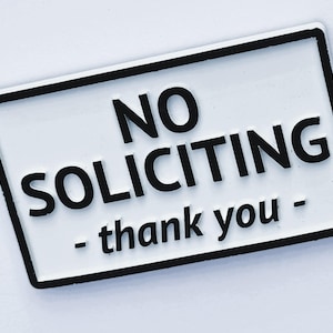 May include: White rectangular sign with black border and text "NO SOLICITING - thank you" in bold black letters. The sign is designed to discourage unwanted sales pitches.