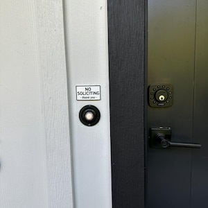 May include: A close-up of a front door with a black door handle and a black doorbell. A small white sign with the words "NO SOLICITING - thank you" is affixed to the door.