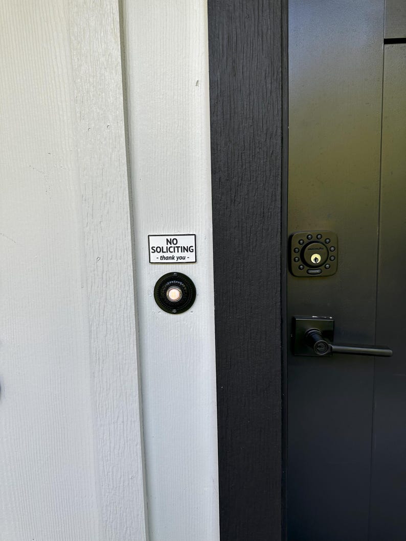 May include: A close-up of a front door with a black door handle and a black doorbell. A small white sign with the words "NO SOLICITING - thank you" is affixed to the door.
