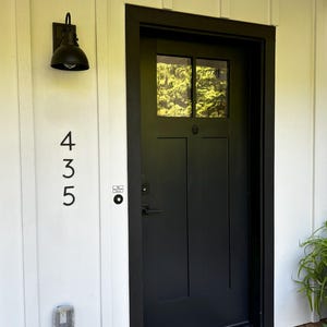 May include: Black front door with a window, set in a black frame. The number 435 is displayed vertically on the white siding to the left of the door. A black outdoor light fixture is mounted above the numbers.