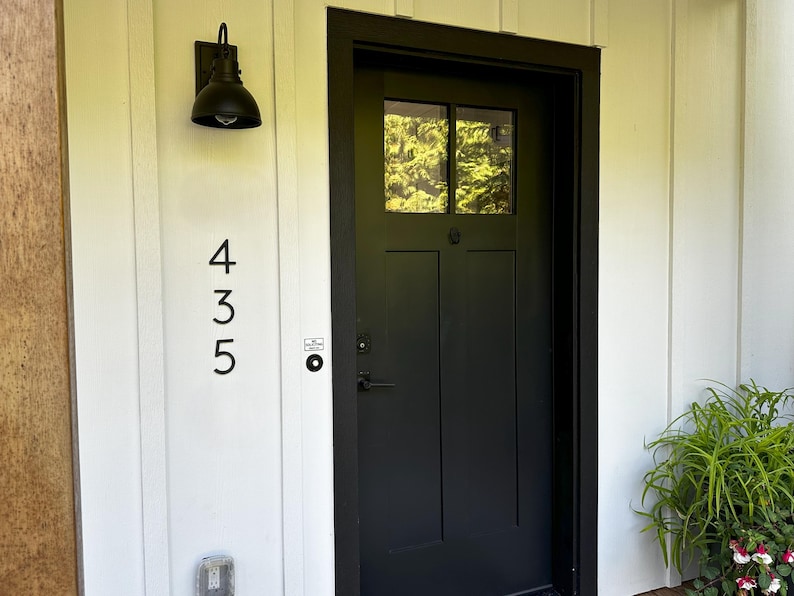 May include: Black front door with a window, set in a black frame. The number 435 is displayed vertically on the white siding to the left of the door. A black outdoor light fixture is mounted above the numbers.