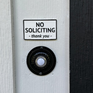 May include: A white door with a black and white "No Soliciting" sign and a black doorbell. The sign has black text that reads "NO SOLICITING - thank you -" in a rectangular frame. The doorbell is round with a white button.