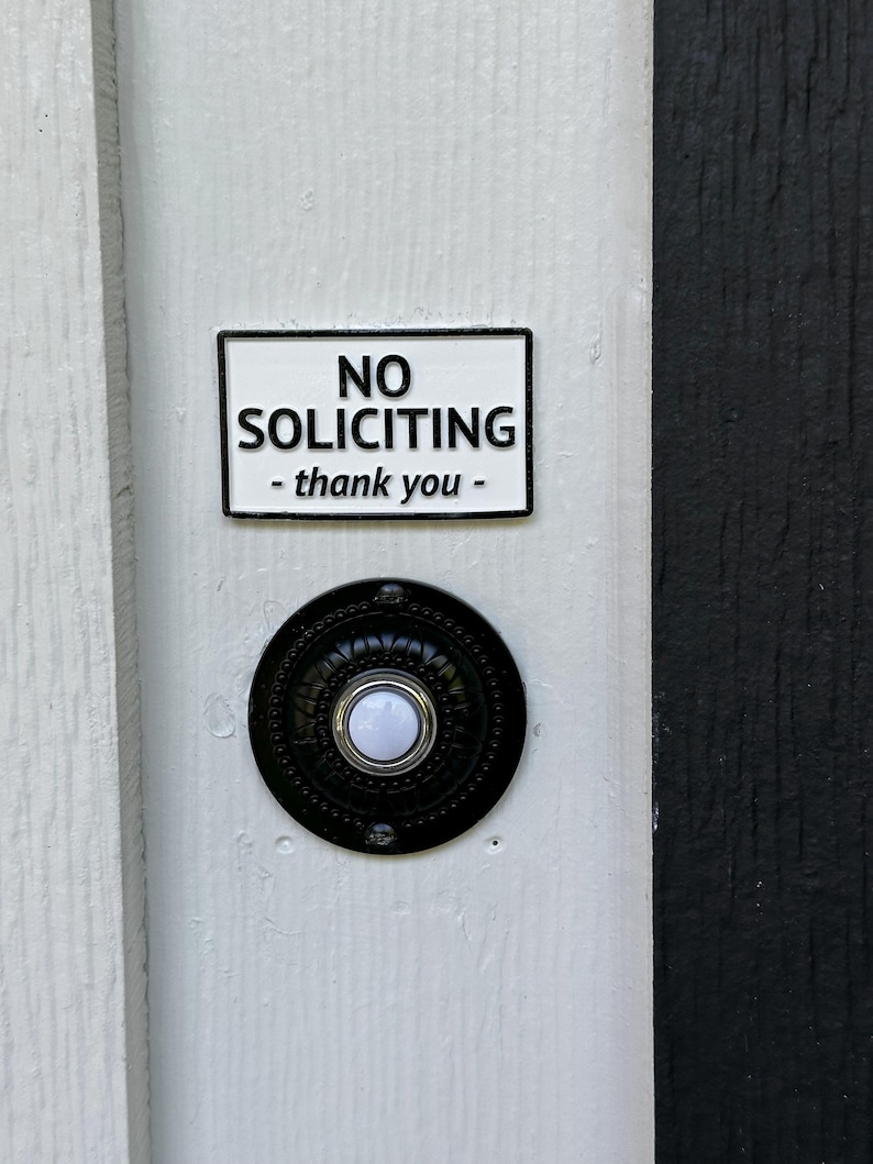 May include: A white door with a black and white "No Soliciting" sign and a black doorbell. The sign has black text that reads "NO SOLICITING - thank you -" in a rectangular frame. The doorbell is round with a white button.