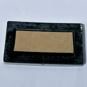 May include: A rectangular black frame with a light brown insert. The frame has a slightly textured surface and a glossy finish. The insert is a lighter shade of brown and appears to be made of cardboard or paper.