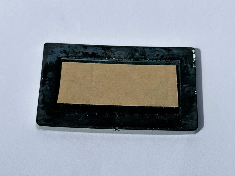 May include: A rectangular black frame with a light brown insert. The frame has a slightly textured surface and a glossy finish. The insert is a lighter shade of brown and appears to be made of cardboard or paper.