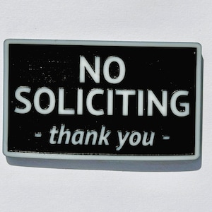 May include: A rectangular black and white sign with the words "NO SOLICITING - thank you -" in white lettering. The sign has a white border and is set against a white background.