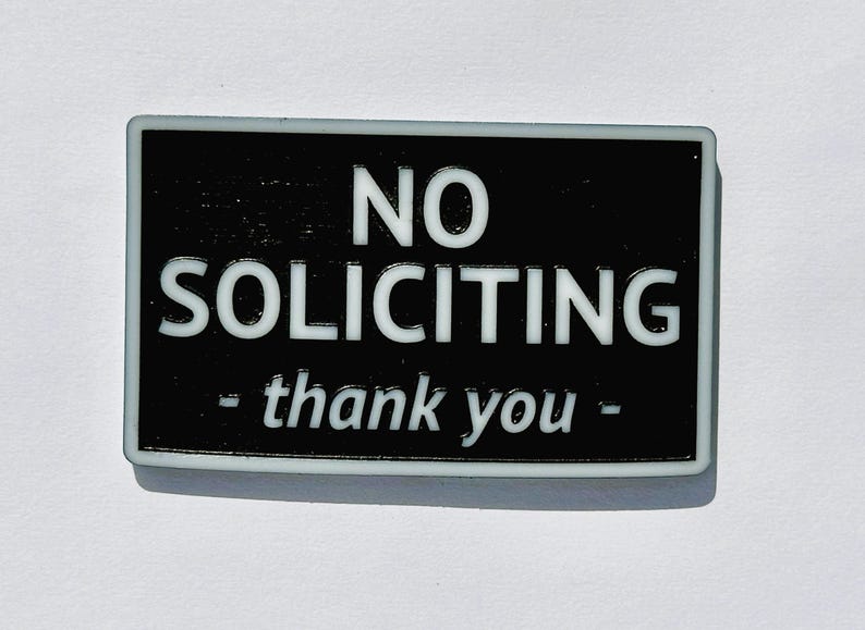 May include: A rectangular black and white sign with the words "NO SOLICITING - thank you -" in white lettering. The sign has a white border and is set against a white background.