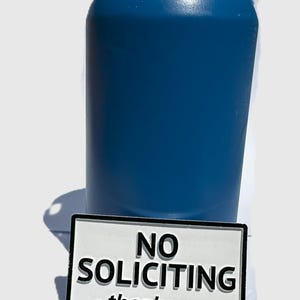May include: A blue insulated tumbler with a black rim and a white rectangular sign that reads "NO SOLICITING - thank you -" in black text. The tumbler is cylindrical and appears to be made of metal.