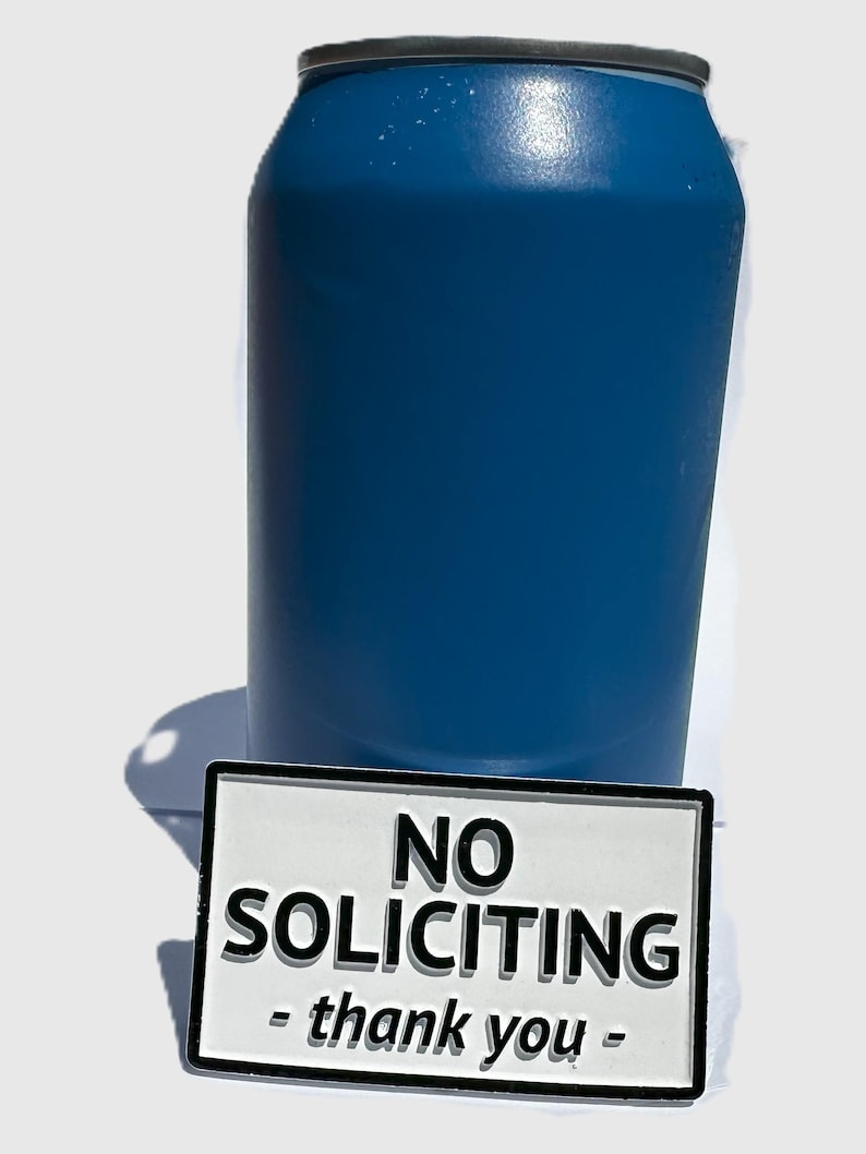 May include: A blue insulated tumbler with a black rim and a white rectangular sign that reads "NO SOLICITING - thank you -" in black text. The tumbler is cylindrical and appears to be made of metal.
