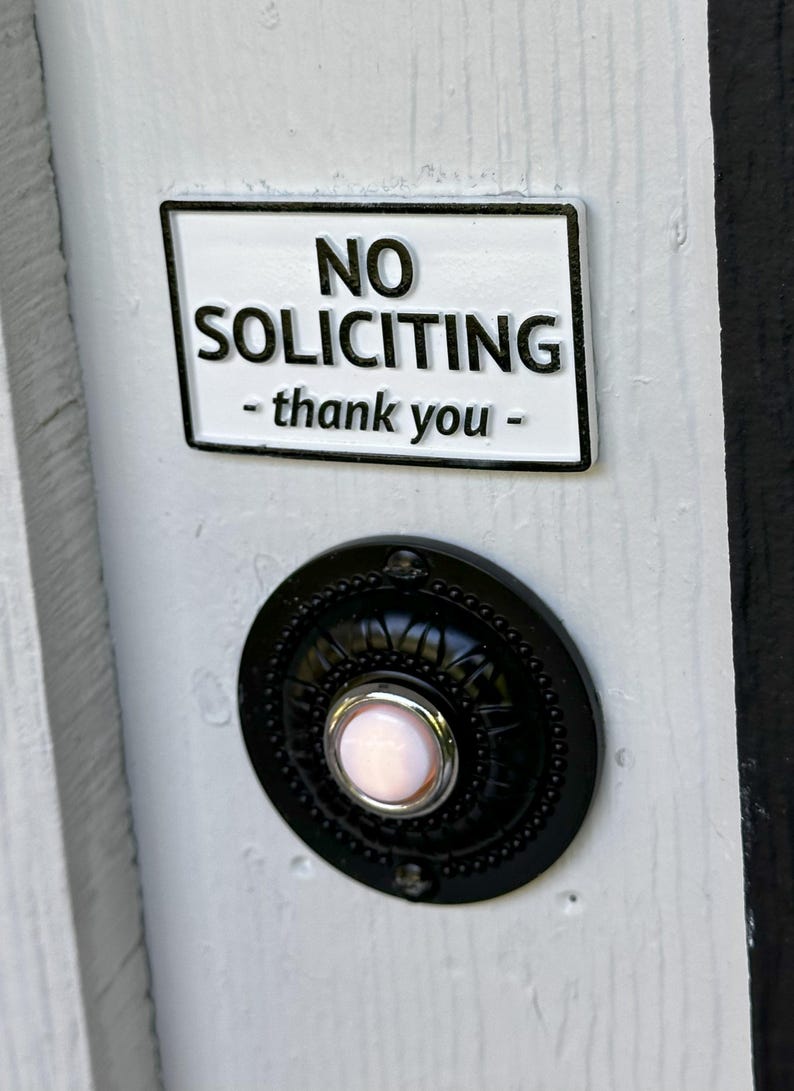 May include: A white rectangular sign with a black border that reads 'NO SOLICITING - thank you -'. Below the sign is a black, round doorbell with a pink illuminated button in the center. The doorbell is mounted on a white surface.