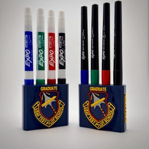May include: Two blue pen holders with the text "GRADUATE" and a patch that reads "USAF TEST PILOT SCHOOL". Each holder contains four pens. One holder has blue, green, red, and black Expo dry erase markers. The other has black wet erase markers.